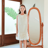 A young girl stands indoors in front of a mirror, wearing the KIKI and JOJO Girls Elegance A-Line Chiffon Trim Dress in champagne, paired with white socks and shoes. Green leaves are visible through the window behind her.