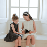 Two young girls sit on a window seat, one wearing a dark dress and the other in the KIKI and JOJO Girls Elegance A-Line Chiffon Trim Dress in Champagne, gently holding and petting a small black and white puppy.