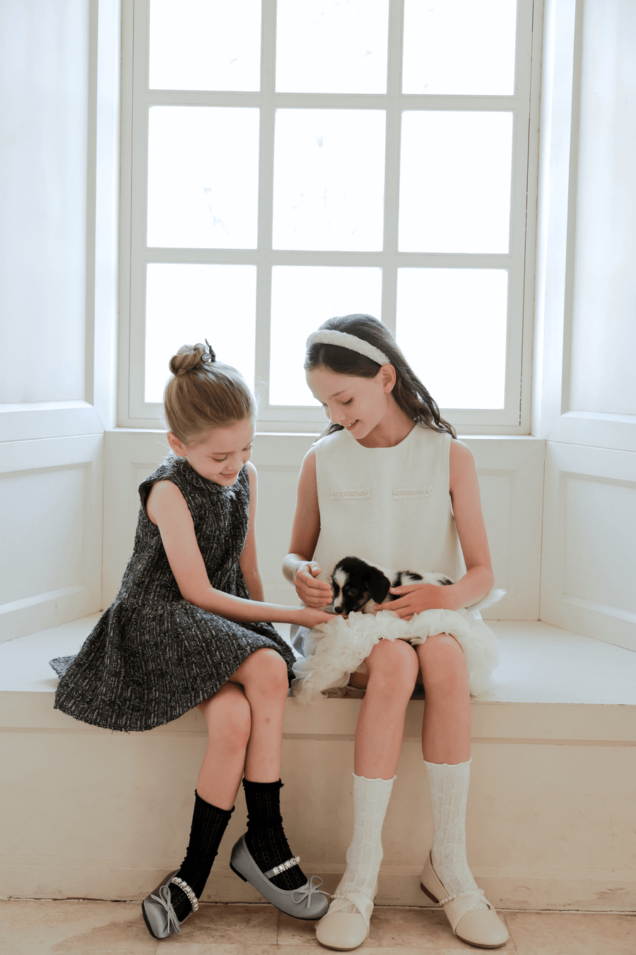 Two young girls sit on a window seat, one wearing a dark dress and the other in the KIKI and JOJO Girls Elegance A-Line Chiffon Trim Dress in Champagne, gently holding and petting a small black and white puppy.