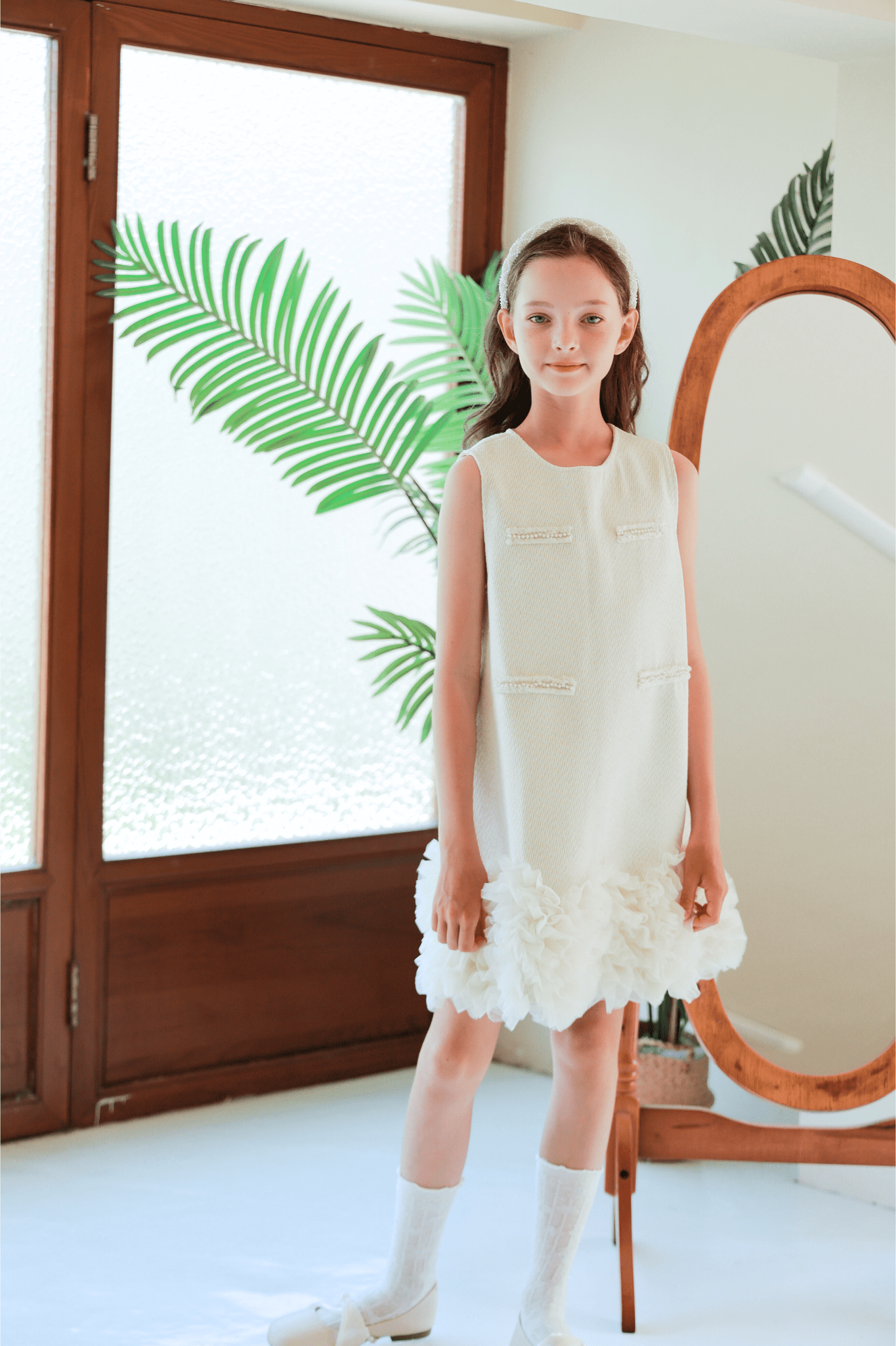 Indoors, a young girl wears the KIKI and JOJO Girls Elegance A-Line Chiffon Trim Dress in champagne, styled with white shoes and a headband. A large leaf and mirror appear in the background.