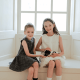 Two young girls sit on a windowsill, one wearing the KIKI and JOJO Girls Elegance A-Line Chiffon Trim Dress in Champagne, the other in white. Together, they gently hold a small black and white puppy.
