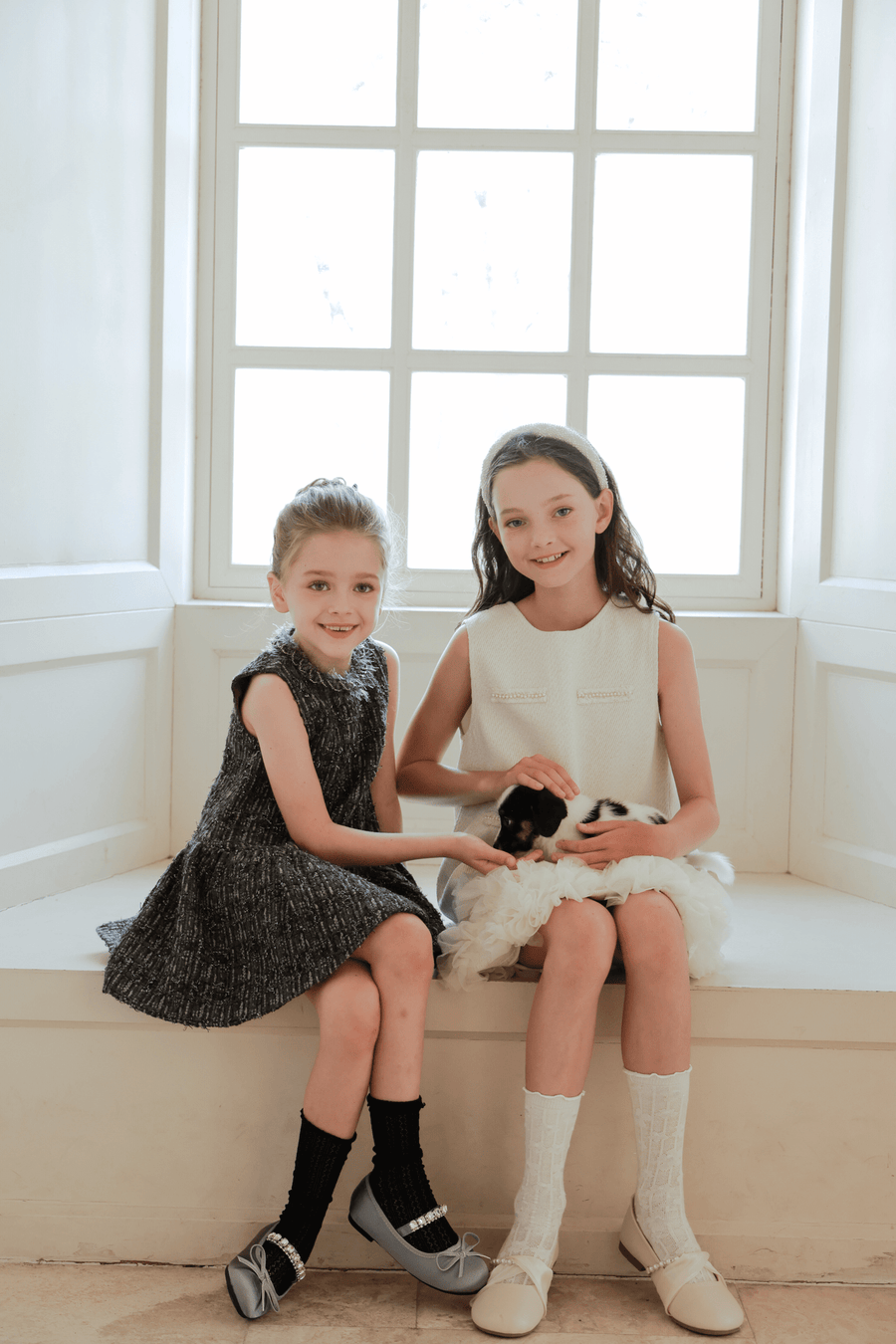 Two young girls sit on a windowsill, one wearing the KIKI and JOJO Girls Elegance A-Line Chiffon Trim Dress in Champagne, the other in white. Together, they gently hold a small black and white puppy.