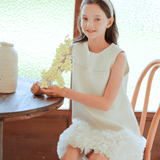 A girl wears the KIKI and JOJO Girls Elegance A-Line Chiffon Trim Dress in champagne, sitting on a wooden chair by a table with yellow flowers, a ceramic vase nearby, and a textured glass window behind her.