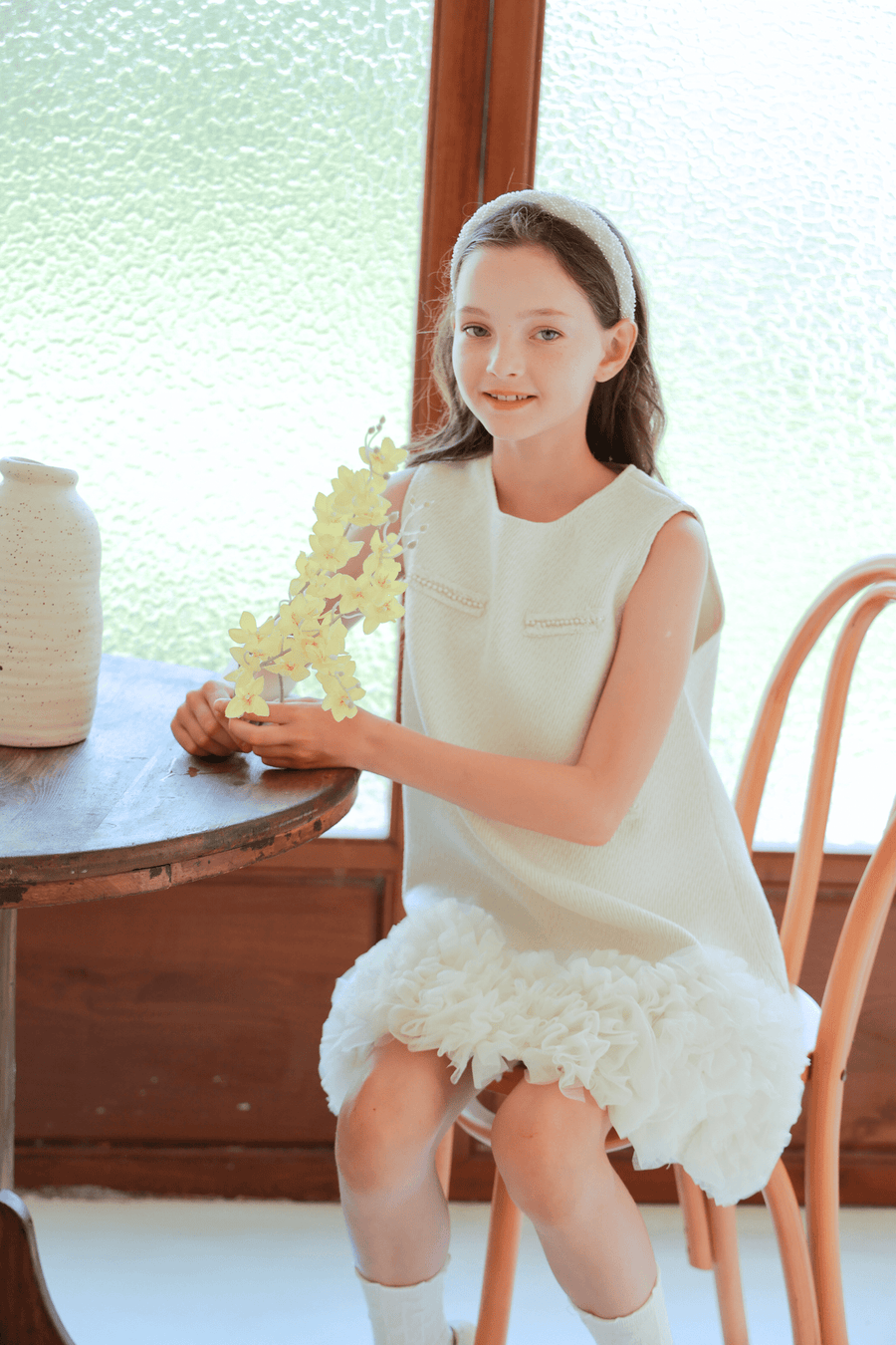 A girl wears the KIKI and JOJO Girls Elegance A-Line Chiffon Trim Dress in champagne, sitting on a wooden chair by a table with yellow flowers, a ceramic vase nearby, and a textured glass window behind her.