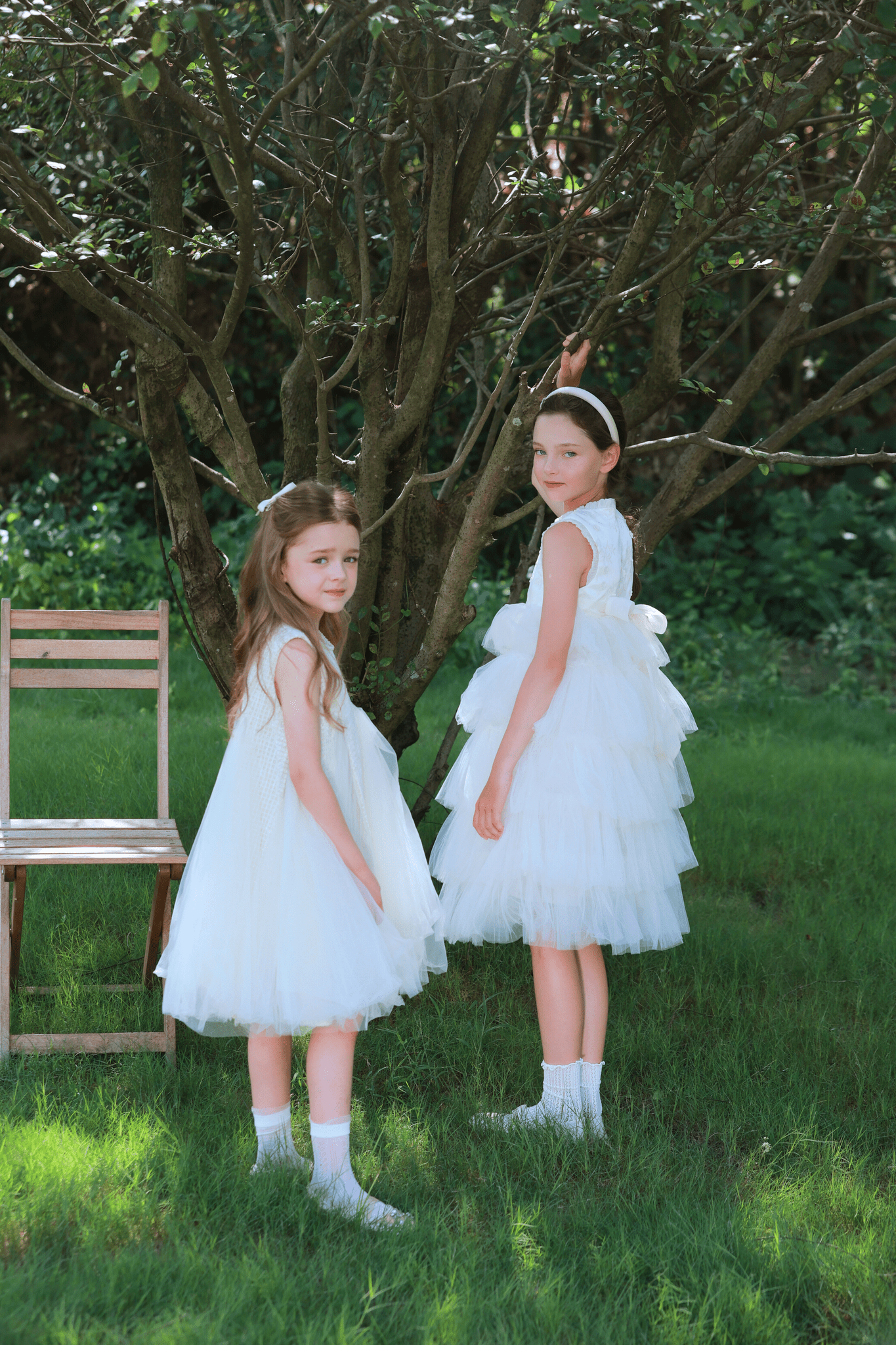 Two young girls in KIKI and JOJO's Girls Embroidered Satin & Tulle Dress - Ivory stand on grass near a wooden chair and tree; one faces forward while the other makes a peace sign—a perfect special occasion moment.