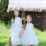 Two young girls wear KIKI and JOJO’s Girls Embroidered Satin & Tulle Dress in ivory while walking hand-in-hand on grass, with a rustic hut and greenery behind them. One carries a bouquet—ideal for special occasions.