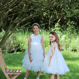 Two girls wear the KIKI and JOJO Girls Embroidered Satin & Tulle Dress in ivory, standing on grass under a tree beside a wooden chair, with a lush garden backdrop—perfect for special occasions.