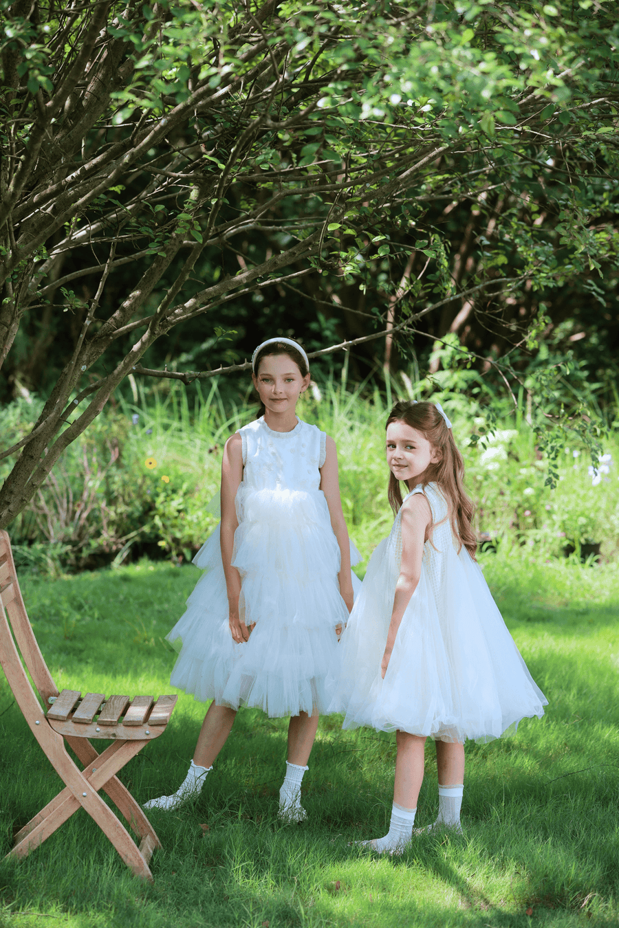 Two girls wear the KIKI and JOJO Girls Embroidered Satin & Tulle Dress in ivory, standing on grass under a tree beside a wooden chair, with a lush garden backdrop—perfect for special occasions.