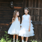 Two young girls in KIKI and JOJO Girls Embroidered Satin & Tulle Dress - Ivory stand side by side before a rustic wooden building with a thatched roof and lantern, perfect for special occasions.