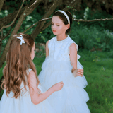 Two young girls wear KIKI and JOJO Girls Embroidered Satin & Tulle Dresses in ivory, standing together under a sunlit tree—ideal for special occasions.