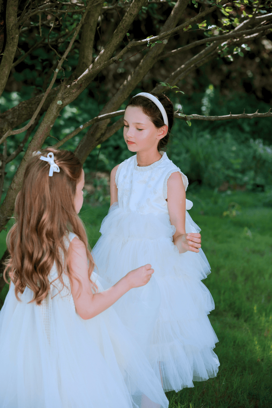 Two young girls wear KIKI and JOJO Girls Embroidered Satin & Tulle Dresses in ivory, standing together under a sunlit tree—ideal for special occasions.