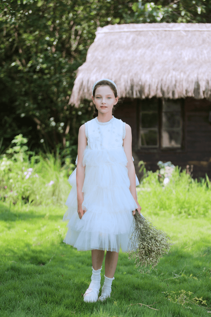 A young girl wears the KIKI and JOJO Girls Embroidered Satin & Tulle Dress in ivory, standing on grass with a dried flower bouquet and a thatched-roof cottage behind—ideal for special occasions.