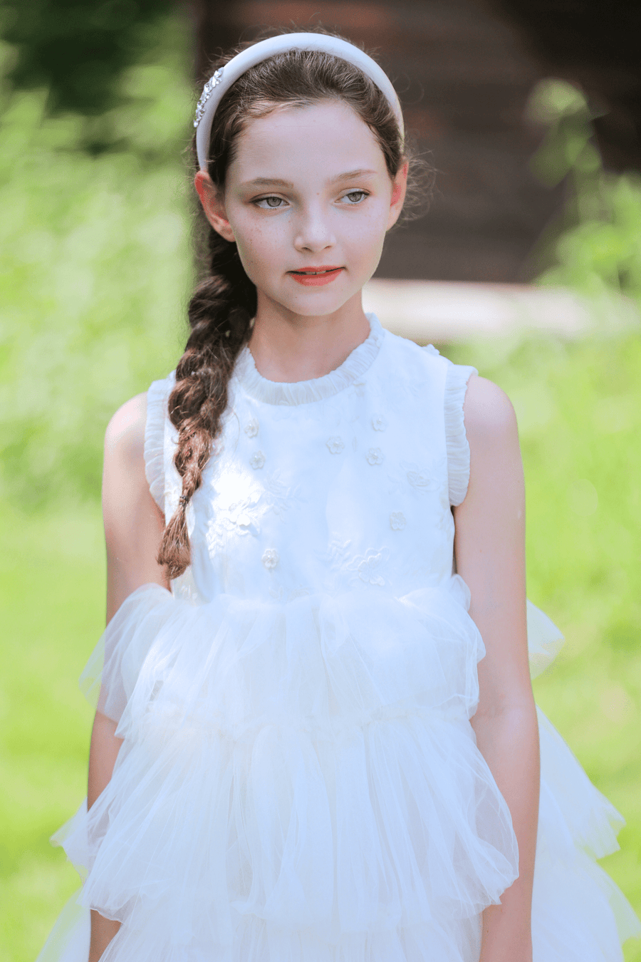 A young girl with a braid and headband wears the KIKI and JOJO Girls Embroidered Satin & Tulle Dress in ivory, ideal for special occasions, standing outdoors in natural light.