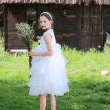 A young girl wears the KIKI and JOJO Girls Embroidered Satin & Tulle Dress in ivory with a headband, holding flowers on grass before a thatched wooden hut—perfect for special occasions.