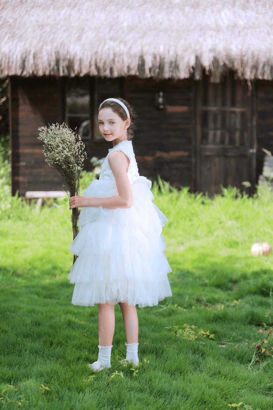 A young girl wears the KIKI and JOJO Girls Embroidered Satin & Tulle Dress in ivory with a headband, holding flowers on grass before a thatched wooden hut—perfect for special occasions.