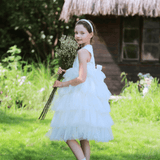A young girl in a KIKI and JOJO Girls Embroidered Satin & Tulle Dress - Ivory stands on grass with a headband and dried flower bouquet, framed by a thatched-roof cottage—an ideal look for special occasions.