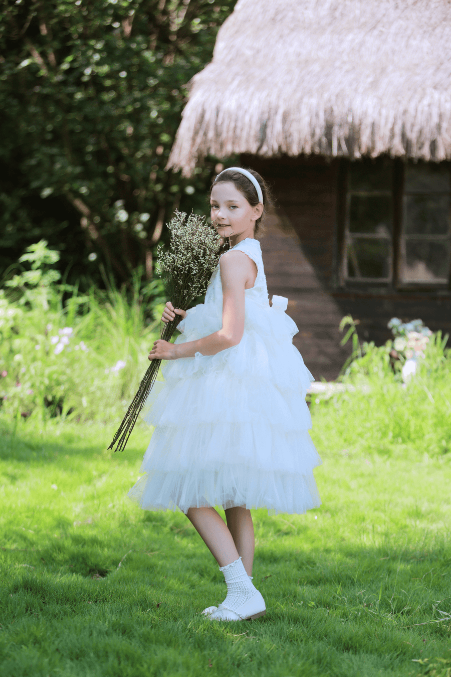 A young girl in a KIKI and JOJO Girls Embroidered Satin & Tulle Dress - Ivory stands on grass with a headband and dried flower bouquet, framed by a thatched-roof cottage—an ideal look for special occasions.