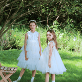 Two girls wear KIKI and JOJO Girls Embroidered Satin & Tulle Dress in ivory, standing on grass under a tree near a wooden chair. The lush green backdrop creates an ideal setting for special occasions.