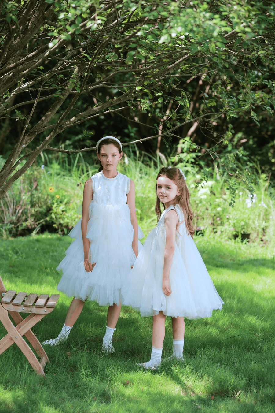 Two girls wear KIKI and JOJO Girls Embroidered Satin & Tulle Dress in ivory, standing on grass under a tree near a wooden chair. The lush green backdrop creates an ideal setting for special occasions.