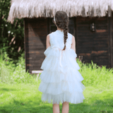 A young girl in a KIKI and JOJO Girls Embroidered Satin & Tulle Dress - Ivory stands on grass facing a wooden house with a thatched roof, her hair in a braid—perfect for special occasions.