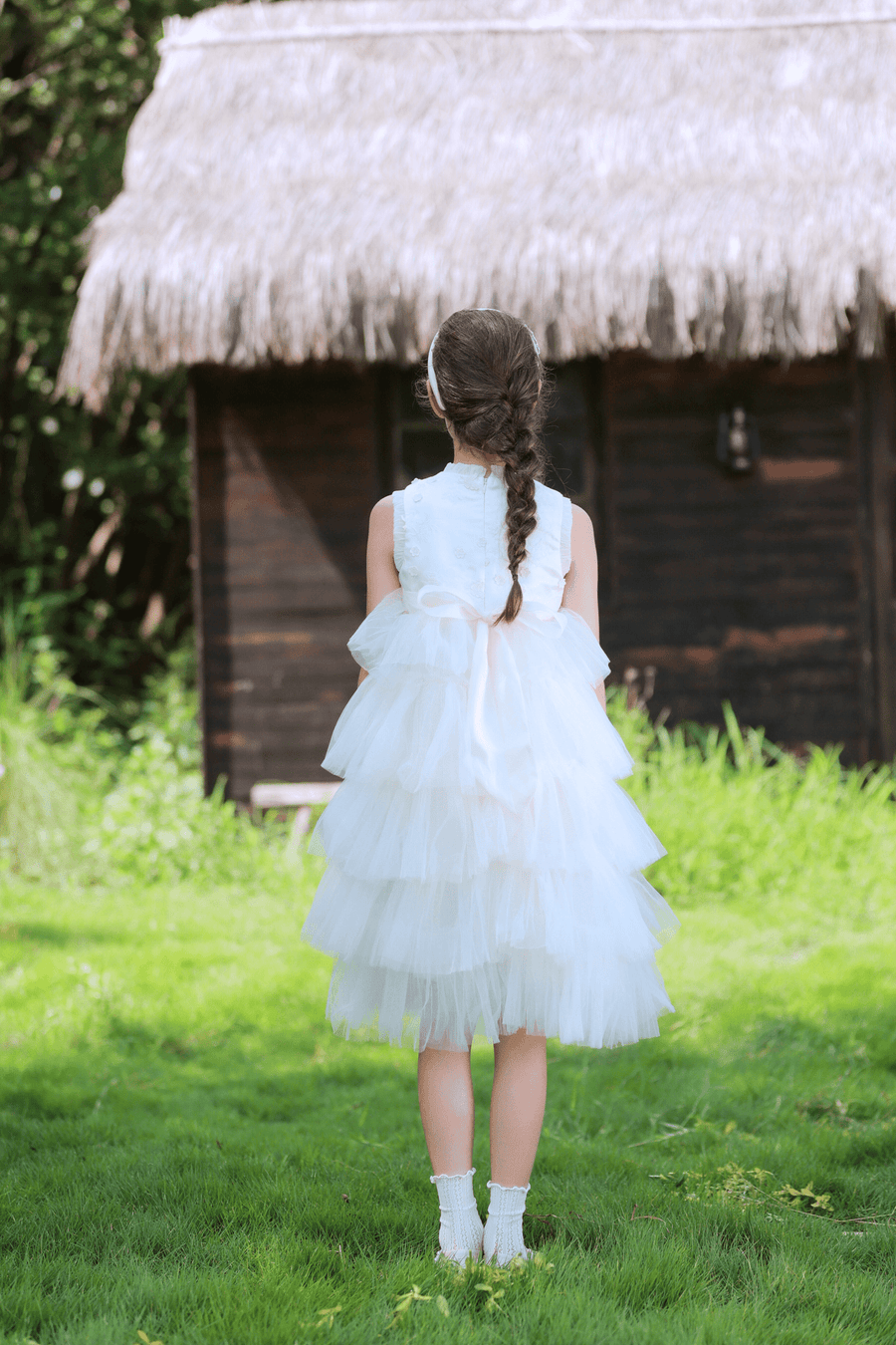 A young girl in a KIKI and JOJO Girls Embroidered Satin & Tulle Dress - Ivory stands on grass facing a wooden house with a thatched roof, her hair in a braid—perfect for special occasions.