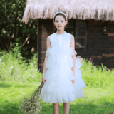 A girl in a KIKI and JOJO Girls Embroidered Satin & Tulle Dress - Ivory stands on grass with a bouquet of dried flowers, a thatched-roof wooden house behind her—perfect for special occasions.