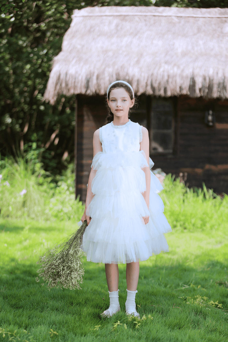 A girl in a KIKI and JOJO Girls Embroidered Satin & Tulle Dress - Ivory stands on grass with a bouquet of dried flowers, a thatched-roof wooden house behind her—perfect for special occasions.