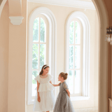 Two girls in KIKI and JOJO Girls Pearl-Embroidered Satin Dress - Pure White stand in a bright hallway with arched windows and cream-colored walls.