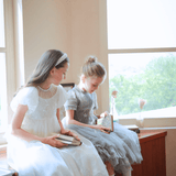 Two young girls in KIKI and JOJO Girls Pearl-Embroidered Satin Dresses in pure white sit by a window on a wooden surface, each holding a book as sunlight glows on flower vases along the windowsill.
