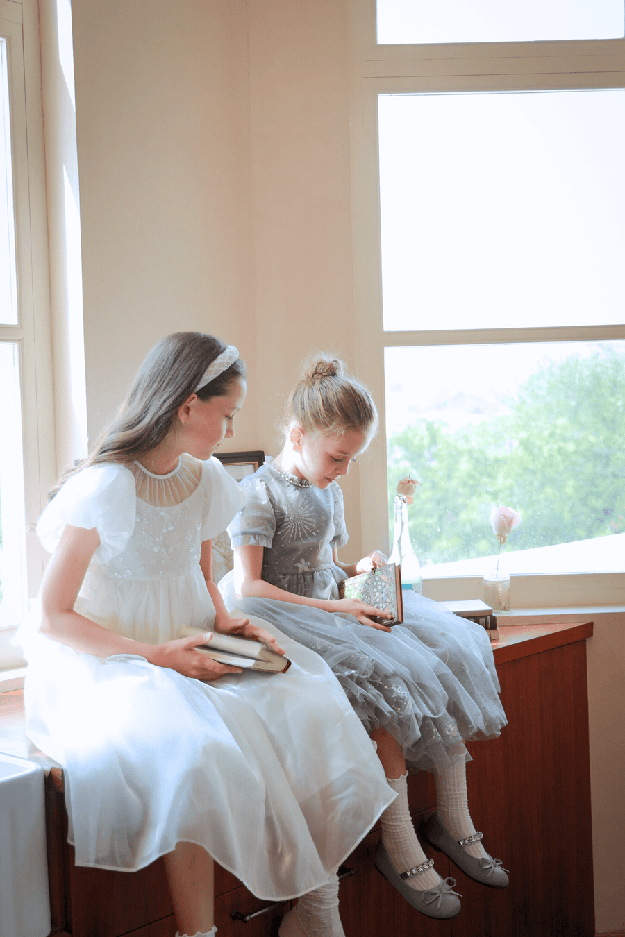 Two young girls in KIKI and JOJO Girls Pearl-Embroidered Satin Dresses in pure white sit by a window on a wooden surface, each holding a book as sunlight glows on flower vases along the windowsill.