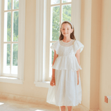 A young girl wears the KIKI and JOJO Girls Pearl-Embroidered Satin Dress in Pure White, standing indoors by tall windows as sunlight highlights the delicate pearl embroidery on her gown.