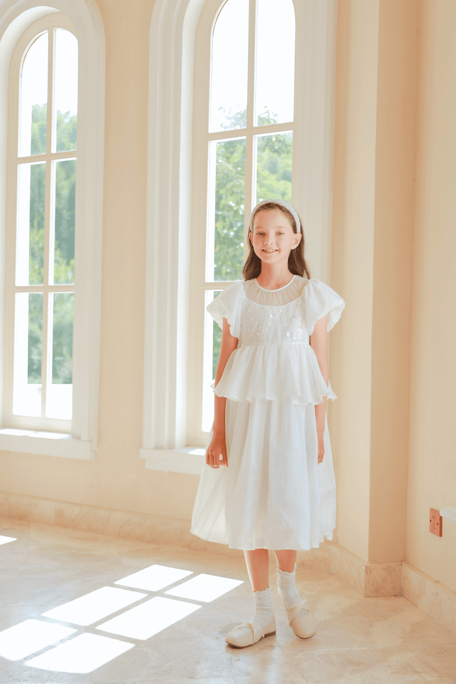 A young girl wears the KIKI and JOJO Girls Pearl-Embroidered Satin Dress in Pure White, standing indoors by tall windows as sunlight highlights the delicate pearl embroidery on her gown.