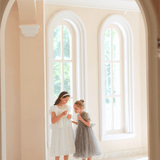 Two young girls stand in a bright, arched-windowed room; one wears the KIKI and JOJO Girls Pearl-Embroidered Satin Dress in Pure White, while the other is dressed in a charming gray dress.