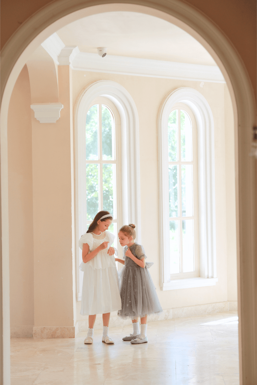 Two young girls stand in a bright, arched-windowed room; one wears the KIKI and JOJO Girls Pearl-Embroidered Satin Dress in Pure White, while the other is dressed in a charming gray dress.