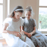Two young girls wearing KIKI and JOJO Girls Pearl-Embroidered Satin Dresses in Pure White sit by a window reading, with a single-stem flower in a vase on the sill, creating a peaceful scene.