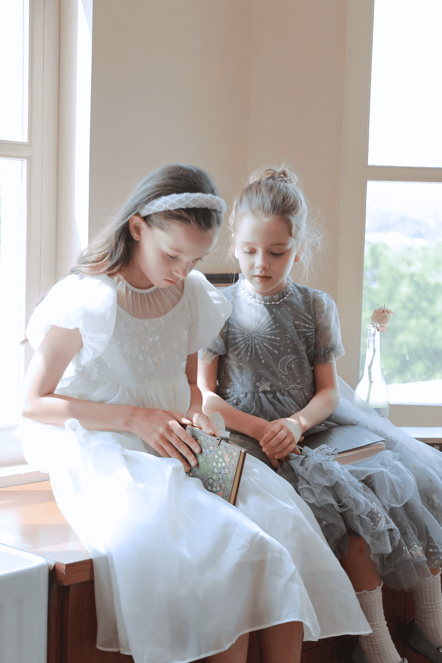 Two young girls wearing KIKI and JOJO Girls Pearl-Embroidered Satin Dresses in Pure White sit by a window reading, with a single-stem flower in a vase on the sill, creating a peaceful scene.