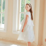 A girl in the KIKI and JOJO Girls Pearl-Embroidered Satin Dress - Pure White stands indoors by tall windows, glancing over her shoulder at the camera as sunlight streams across the floor.