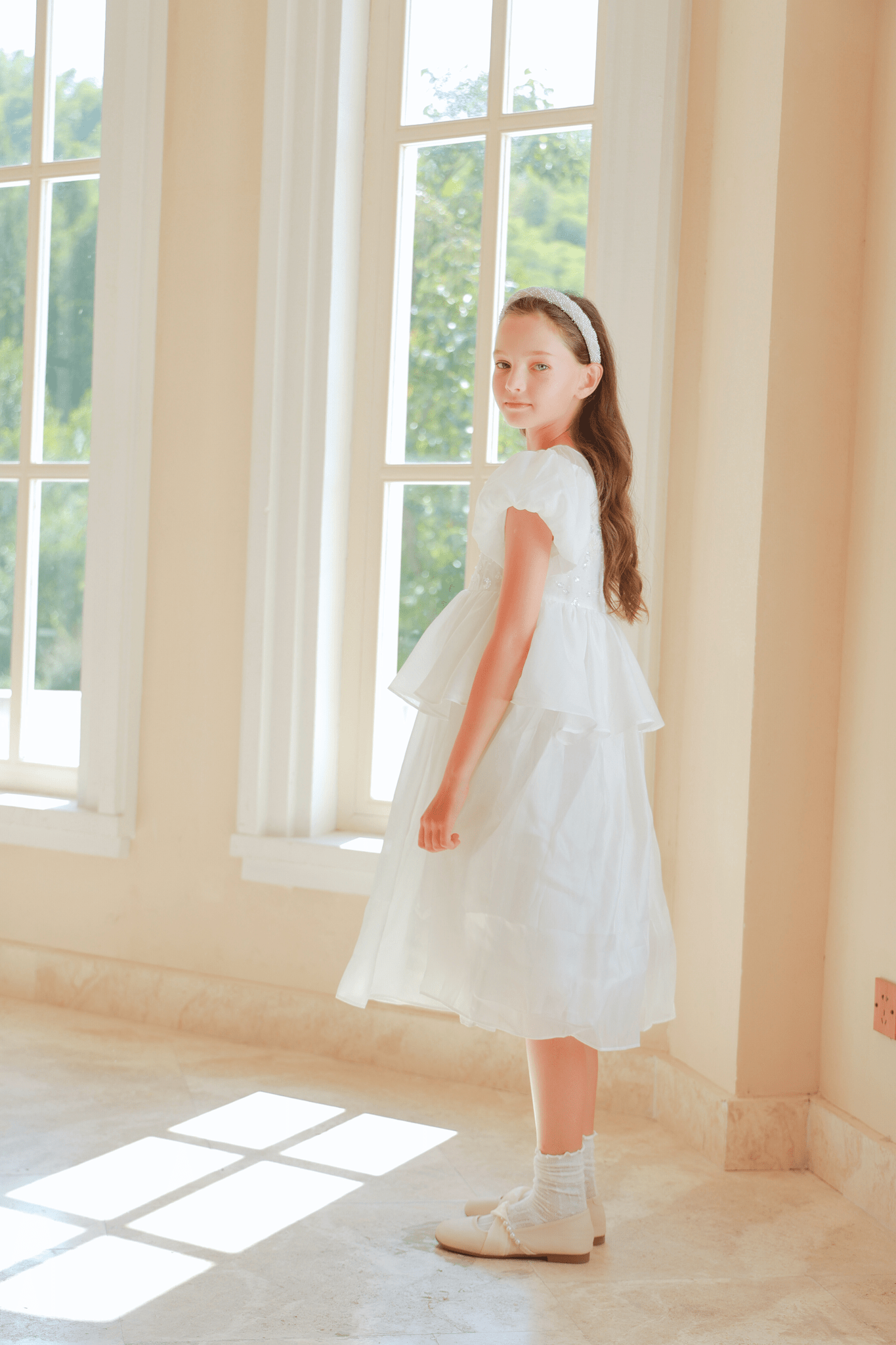 A girl in the KIKI and JOJO Girls Pearl-Embroidered Satin Dress - Pure White stands indoors by tall windows, glancing over her shoulder at the camera as sunlight streams across the floor.