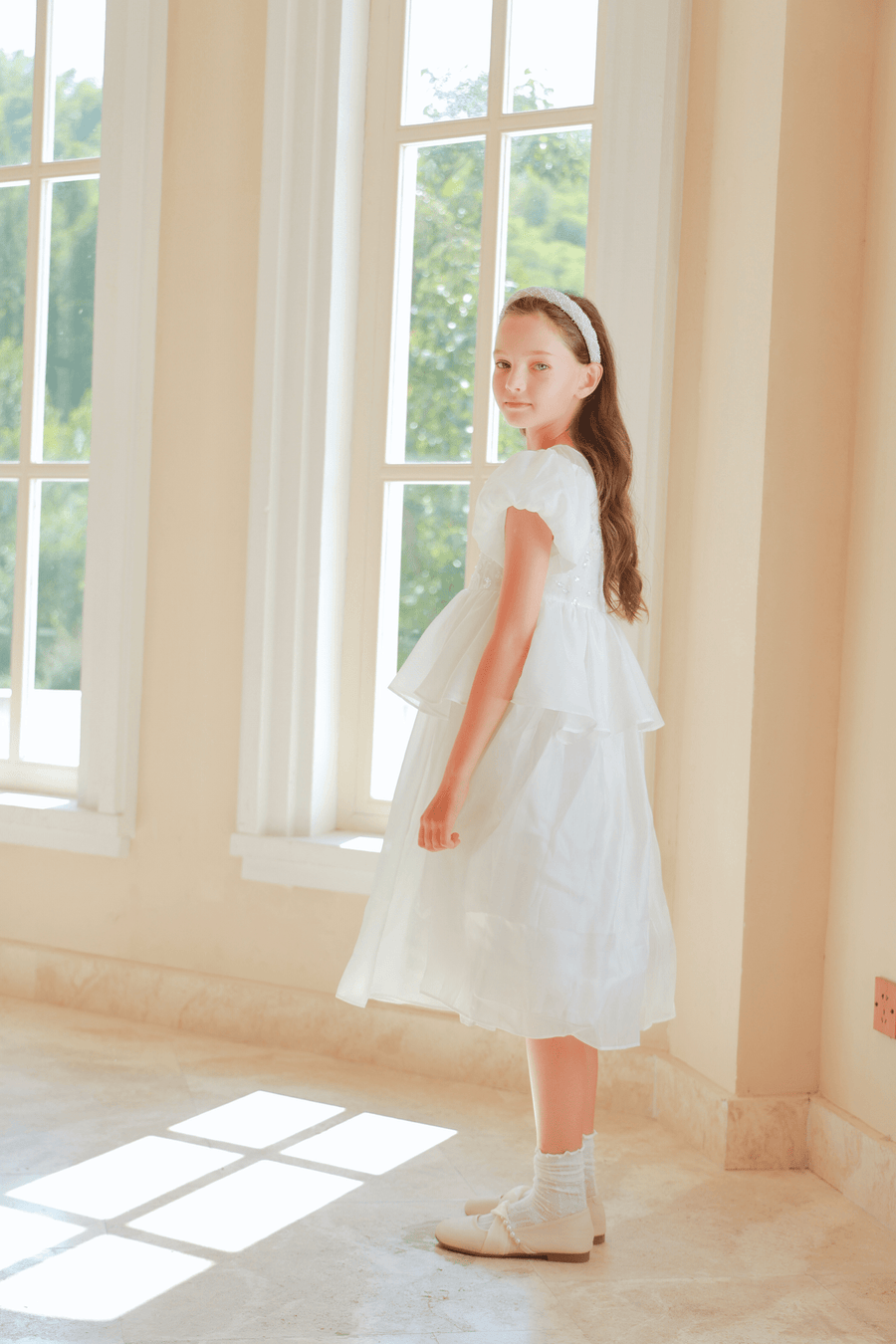 A girl in the KIKI and JOJO Girls Pearl-Embroidered Satin Dress - Pure White stands indoors by tall windows, glancing over her shoulder at the camera as sunlight streams across the floor.