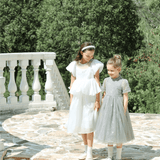 Two young girls in KIKI and JOJO’s Girls Pearl-Embroidered Satin Dress in Pure White stand outdoors on stone steps, surrounded by greenery and a white balustrade.