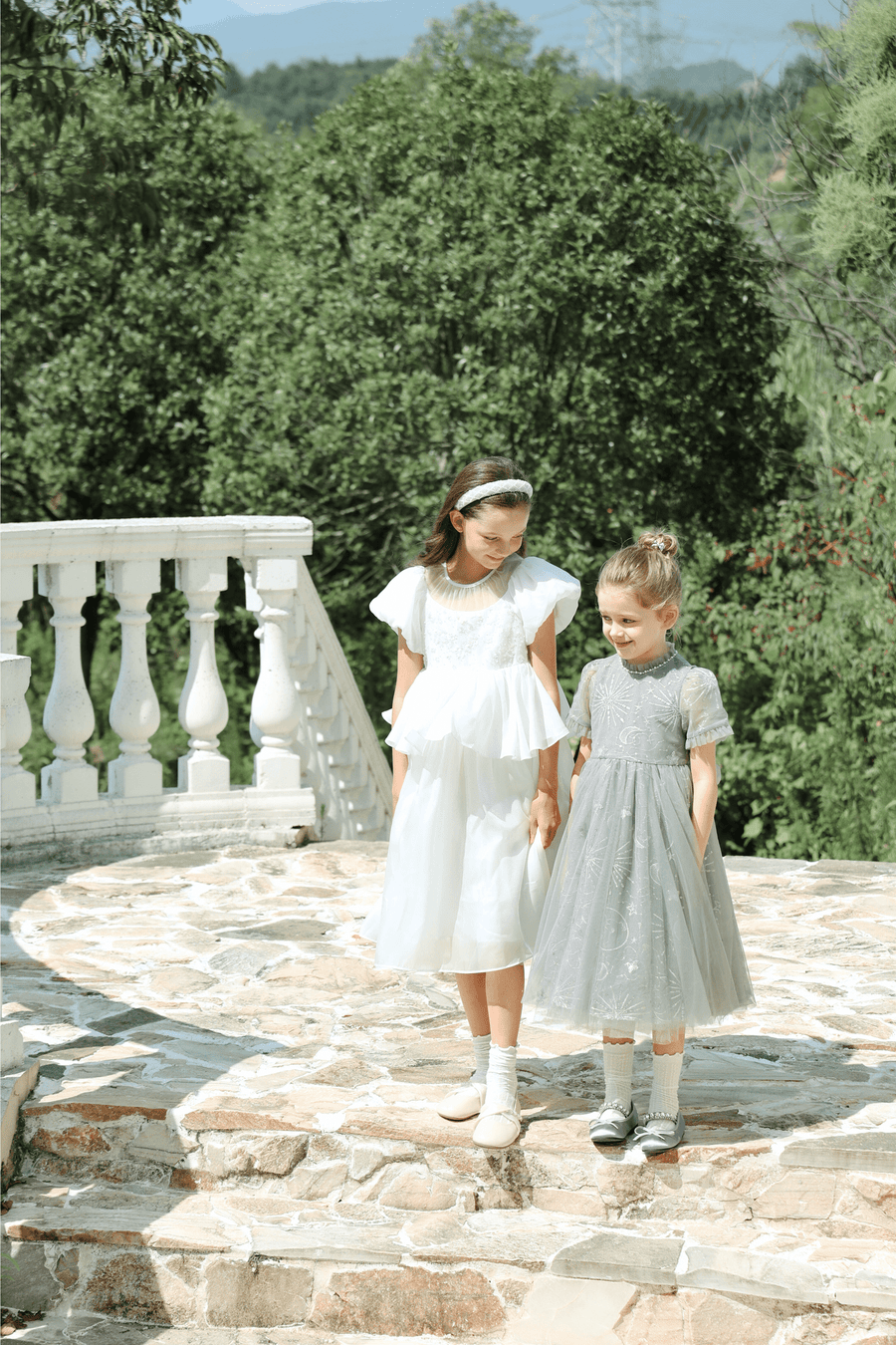 Two young girls in KIKI and JOJO’s Girls Pearl-Embroidered Satin Dress in Pure White stand outdoors on stone steps, surrounded by greenery and a white balustrade.