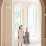 Two young girls stand side by side in a bright room with arched windows, each wearing the KIKI and JOJO Girls Pearl-Embroidered Satin Dress in Pure White, viewed through an arched doorway.