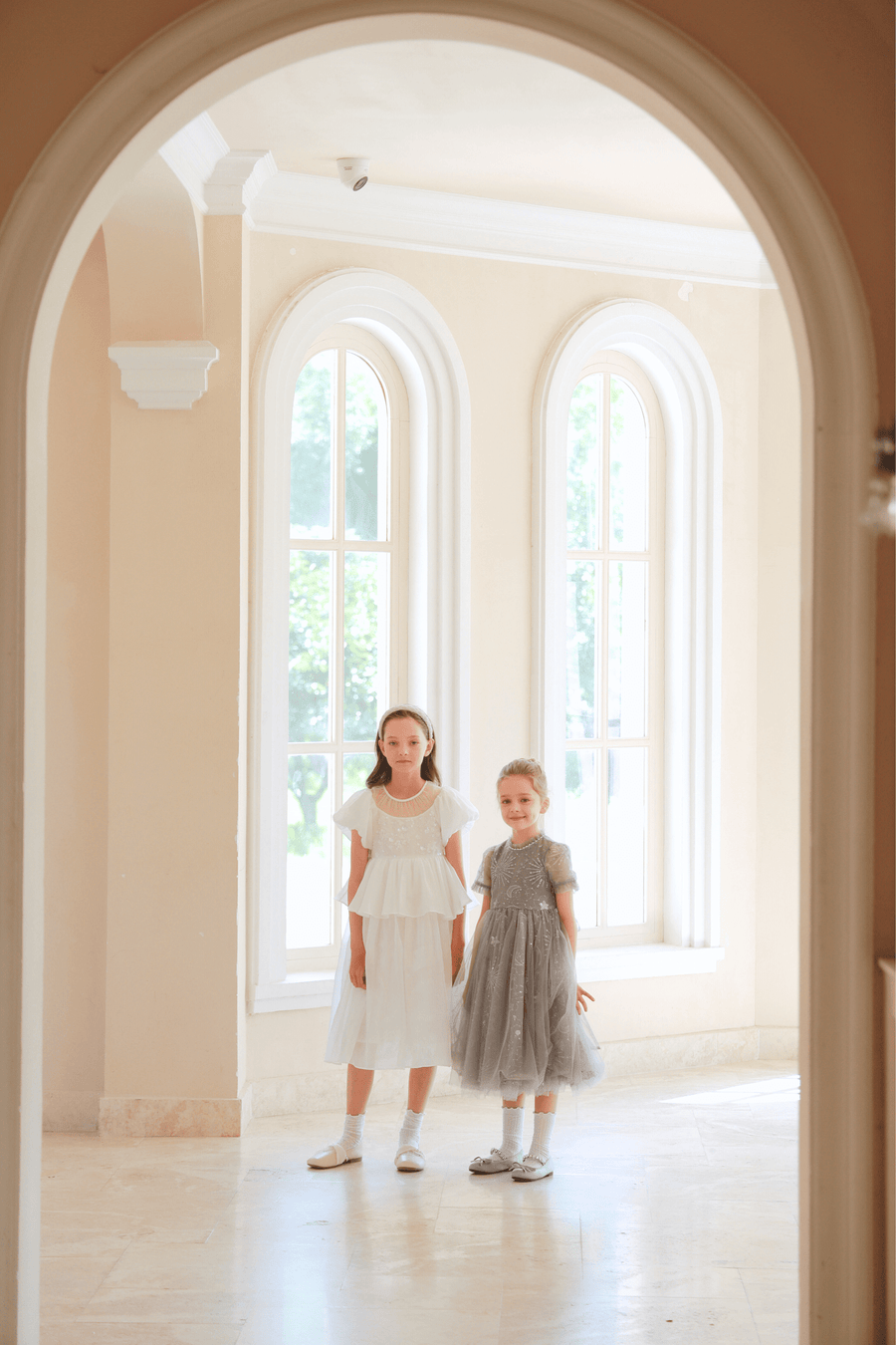 Two young girls stand side by side in a bright room with arched windows, each wearing the KIKI and JOJO Girls Pearl-Embroidered Satin Dress in Pure White, viewed through an arched doorway.