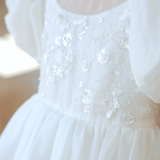Close-up of the KIKI and JOJO Girls Pearl-Embroidered Satin Dress in Pure White, showcasing a sheer neckline, puffy sleeves, floral sequin details, and delicate hand-embroidered pearl accents.