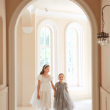 Two young girls in formal dresses, one wearing the KIKI and JOJO Girls Pearl-Embroidered Satin Dress - Pure White, walk hand-in-hand through a bright hallway with large windows in the background.
