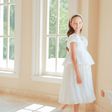 A girl wearing the KIKI and JOJO Girls Pearl-Embroidered Satin Dress in Pure White with a matching headband stands indoors by large sunlit windows.