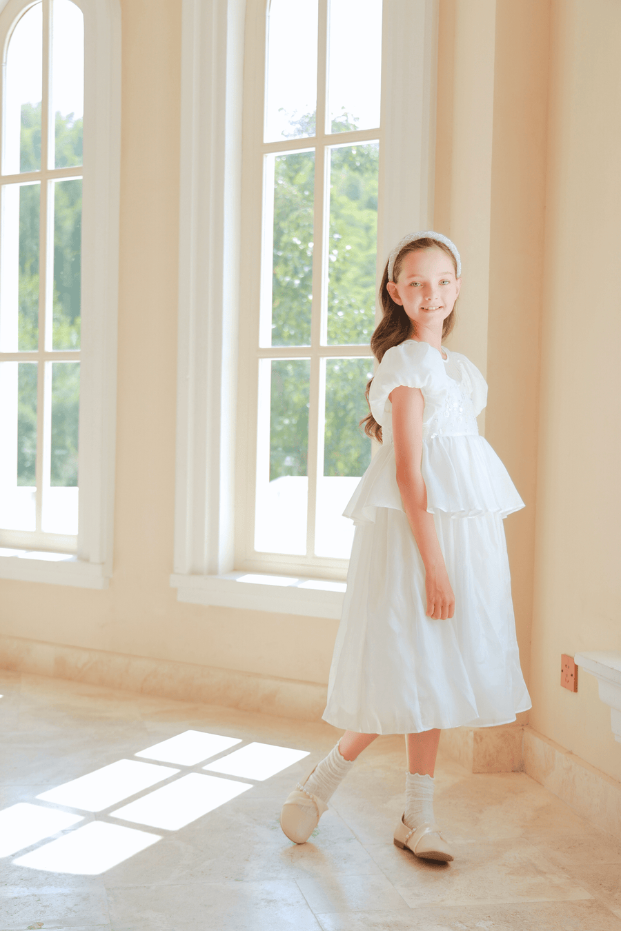 A girl wearing the KIKI and JOJO Girls Pearl-Embroidered Satin Dress in Pure White with a matching headband stands indoors by large sunlit windows.