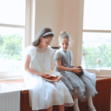 Two young girls in KIKI and JOJO Girls Pearl-Embroidered Satin Dress - Pure White sit on a wooden countertop by large windows, reading books as natural light fills the room.