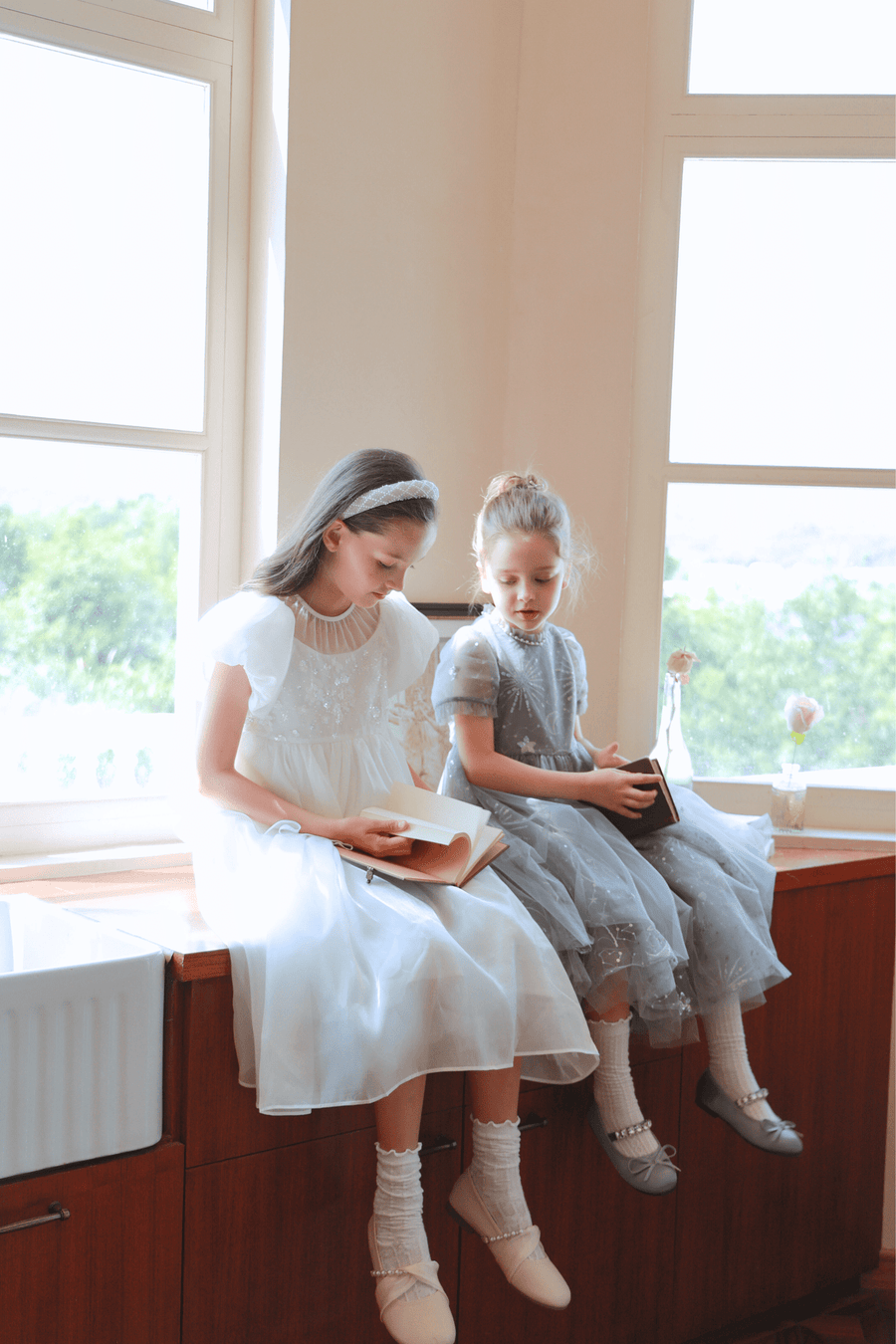 Two young girls in KIKI and JOJO Girls Pearl-Embroidered Satin Dress - Pure White sit on a wooden countertop by large windows, reading books as natural light fills the room.