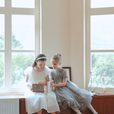 Two young girls in KIKI and JOJO Girls Pearl-Embroidered Satin Dresses in Pure White sit on a wooden cabinet by large windows, admiring a sparkling clutch together.
